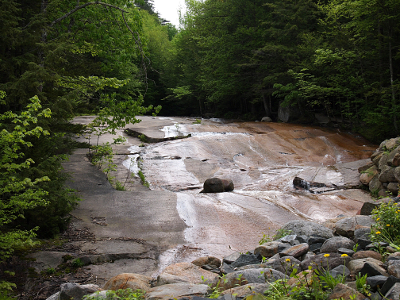 [A large rock surface with many angles and cracks covers a hillside. A number of different paths of water cross the face of the rock.]